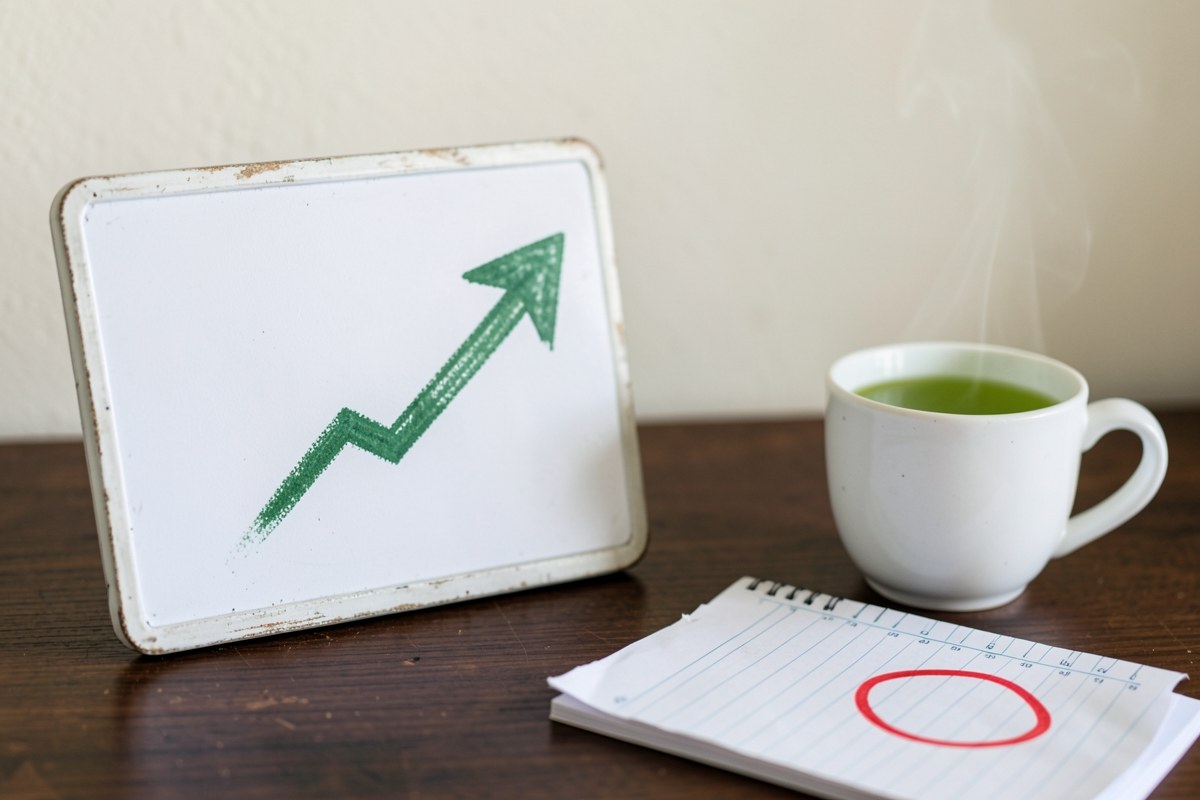 A small whiteboard with a simple financial growth arrow drawn in green, and a calendar with a single red circle on a 15th day, on a desk with a cup of green tea.